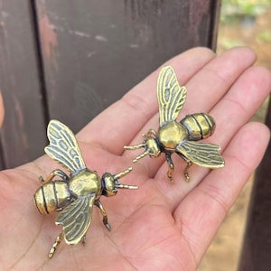 K&ouml;nnte beinhalten: Zwei kleine, detaillierte Messing-Bienenfiguren. Die Bienen haben komplizierte Fl&uuml;gelmuster und sind mit ausgestreckten Beinen dargestellt. Die Figuren werden in der Hand einer Person gehalten, was ihre Gr&ouml;&szlig;e und Handwerkskunst zeigt.