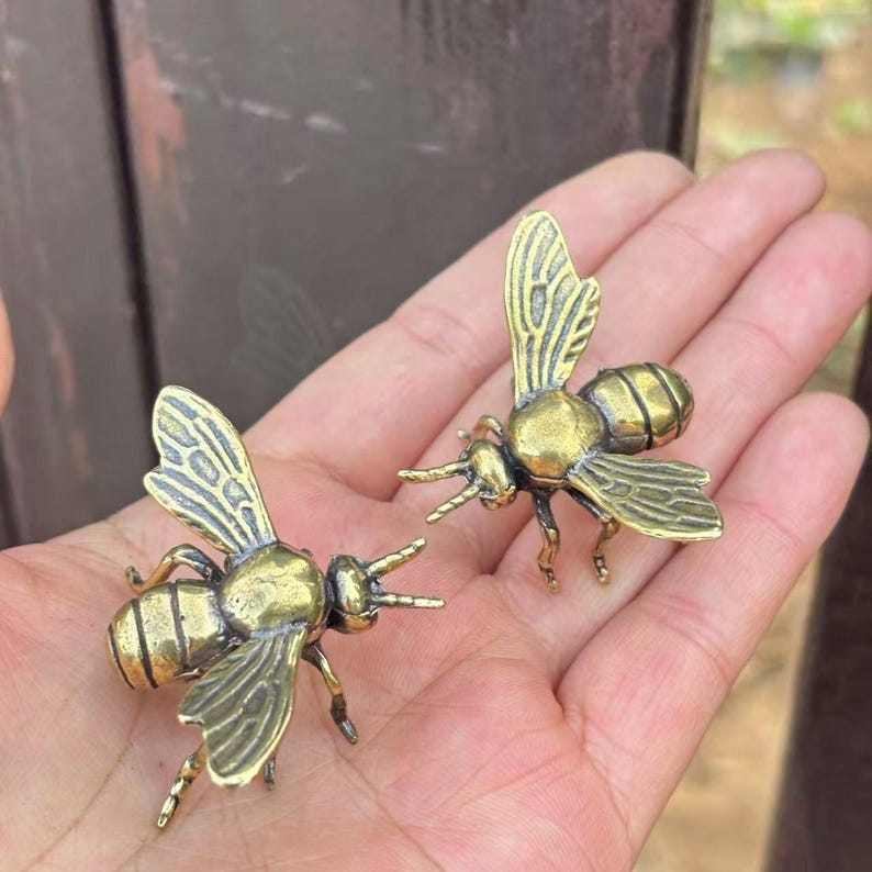 K&ouml;nnte beinhalten: Zwei kleine, detaillierte Messing-Bienenfiguren. Die Bienen haben komplizierte Fl&uuml;gelmuster und sind mit ausgestreckten Beinen dargestellt. Die Figuren werden in der Hand einer Person gehalten, was ihre Gr&ouml;&szlig;e und Handwerkskunst zeigt.