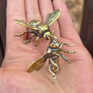 K&ouml;nnte beinhalten: Zwei kleine, detaillierte Messing-Bienenfiguren. Die Bienen sind so positioniert, als w&uuml;rden sie interagieren. Die Fl&uuml;gel und Beine sind sichtbar. Das Messing hat einen goldenen Farbton. Die Figuren sind klein genug, um in eine Handfl&auml;che zu passen.