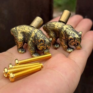 May include: Two brass-toned metal cabinet knobs shaped like standing bears with detailed fur textures. Each knob features a cylindrical mounting post on the back. Four gold-toned metal machine screws are included for installation purposes.