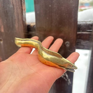 May include: A brass eel figurine with a detailed design. The eel is golden in color and has a textured body, visible gills, and a detailed mouth with tiny teeth. The figurine is held in a hand.