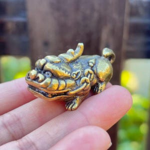 May include: A small, detailed brass figurine of a mythical creature, possibly a Foo Dog or similar guardian figure. The sculpture has intricate features, including a detailed face, paws, and a curled tail. The brass has a warm, golden hue.