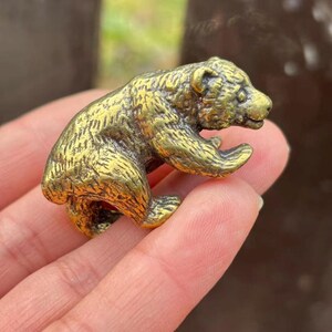 May include: A small, detailed brass bear figurine. The bear is depicted in a crouching position with its paws extended. The brass has a golden hue, and the figurine has intricate detailing, suggesting a handcrafted item.
