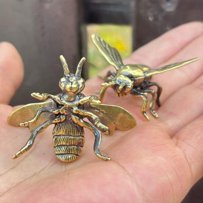 K&ouml;nnte beinhalten: Zwei Bienenfiguren aus Messing. Eine Biene befindet sich in einer Frontalansicht mit ausgebreiteten Fl&uuml;geln und detaillierten K&ouml;rpersegmenten. Die andere Biene ist im Seitenprofil, ebenfalls mit ausgebreiteten Fl&uuml;geln. Beide befinden sich auf neutralem Hintergrund.