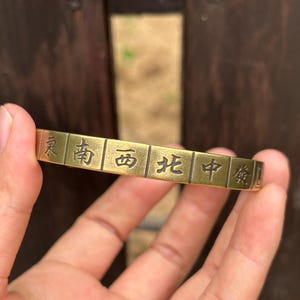 May include: A brass-toned metal cuff bracelet featuring a series of rectangular segments, each engraved with a distinct black Chinese character representing the cardinal directions and Mahjong tiles, including East, South, West, North, and Center.