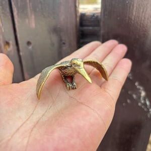 May include: A small, detailed brass bird sculpture with outstretched wings. The bird has a long beak and intricate feather detailing. The sculpture is held in a hand, showcasing its size and craftsmanship. The bird is a decorative item.