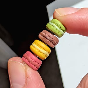 Può includere: Primo piano di cinque macaron in miniatura nei colori rosa, giallo, marrone e verde. I macaron sono tenuti tra due dita, mostrando la loro consistenza dettagliata e il design a strati. Lo sfondo è scuro.