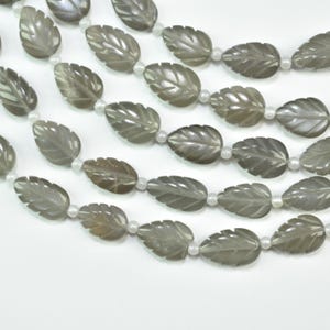 May include: A strand of gray, leaf-shaped beads carved with vein details, alternating with small, round, white spacer beads. The beads are arranged in multiple rows, showcasing a smooth, polished finish and a consistent, repeating pattern.