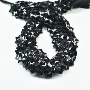 Può includere: Una collana di perline nere sfaccettate di forma geometrica quadrata. Le perline sono infilate insieme, formando un anello, e sono esposte su uno sfondo bianco. Le perline hanno una superficie lucida e riflettente.