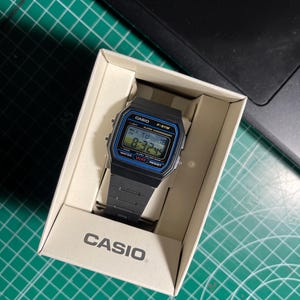 Könnte beinhalten: Eine schwarze Casio F-91W Digitaluhr mit blau-silbernem Display, präsentiert in der Originalverpackung. Die Uhr zeigt die Uhrzeit 8:32:49 an. Auf der Box ist das Casio-Logo abgebildet.