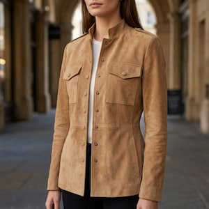 May include: A tan suede jacket featuring a high collar, long sleeves, and a front button closure. The garment includes two chest pockets with buttoned flaps and shoulder epaulets, worn over a plain white shirt and black trousers.
