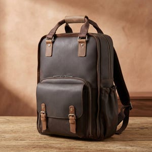 Könnte beinhalten: Ein dunkelbrauner Lederrucksack mit einem Tragegriff, verstellbaren Schultergurten und einer Vordertasche, die mit zwei Schnallenriemen gesichert ist. Die Tasche verfügt über Seitentaschen und Metallbeschläge sowie ein strukturiertes Design mit sichtbaren Nähten.