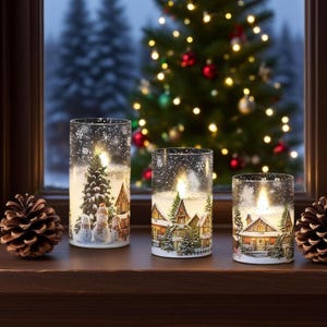 Op de afbeelding: Drie cilindrische glazen kandelaars met winterdorpscènes en LED-verlichting. De houders bevatten illustraties van met sneeuw bedekte huizen, bomen en sneeuwpoppen. Op de achtergrond is een kerstboom met lichtjes te zien. Twee dennenappels liggen op het houten oppervlak.
