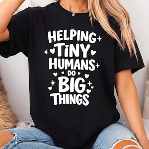 Puede incluir: Camiseta negra con el texto blanco "Helping Tiny Humans Do Big Things". El texto está decorado con corazones y estrellas. La camiseta se usa con vaqueros azules.