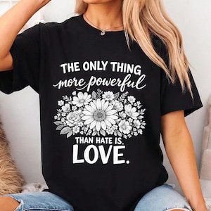 Può includere: T-shirt nero con la scritta bianca "THE ONLY THING MORE POWERFUL THAN HATE IS LOVE" e un motivo floreale. Il disegno presenta una grande margherita circondata da altri fiori e foglie. La maglietta è indossata da una persona che indossa jeans blu.