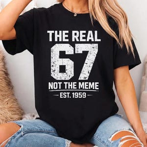 May include: Black t-shirt with white text that reads "THE REAL 67 NOT THE MEME - EST. 1959". The number 67 is large and distressed. The shirt is worn with blue jeans.