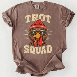 Puede incluir: Una camiseta marrón de manga corta con el gráfico de un pollo que lleva una cinta roja y gafas de sol con patrón de cuadros. El texto TROT SQUAD está impreso en una fuente beige desgastada sobre y debajo de la ilustración.