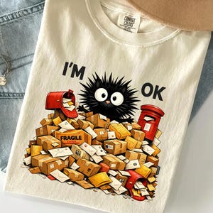 Puede incluir: Camiseta de color marfil con el texto "I'M OK" y un personaje de gato negro de dibujos animados sentado sobre una pila de correo y paquetes. La camiseta se combina con vaqueros azules y un sombrero beige.