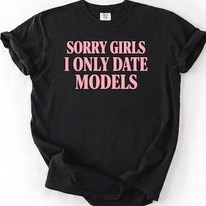 May include: Black t-shirt with the pink text "SORRY GIRLS I ONLY DATE MODELS". The shirt has a crew neck and short sleeves. The text is centered on the front of the shirt.