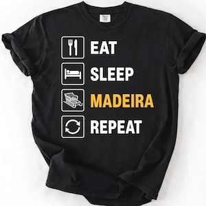 Puede incluir: Una camiseta negra de manga corta con un diseño gráfico blanco de cuatro iconos apilados y texto. El texto dice EAT, SLEEP, MADEIRA en amarillo, y REPEAT. Los iconos representan un tenedor y un cuchillo, una cama, un trineo y una flecha circular.