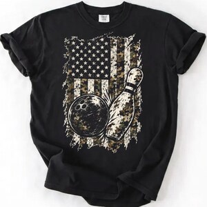 May include: A black short-sleeve t-shirt featuring a graphic print of a bowling ball and a bowling pin centered on the chest. The graphic incorporates a distressed American flag pattern with a camouflage color scheme of green, brown, and tan.