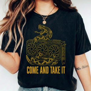 May include: A charcoal grey short-sleeve t-shirt featuring a yellow graphic of a snake coiled around a computer graphics card. The text "COME AND TAKE IT" is printed in yellow capital letters below the illustration of the hardware component.