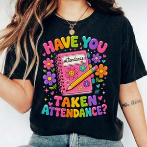 Op de afbeelding: Een zwart t-shirt met korte mouwen en een kleurrijke afbeelding van een roze spiraalblok met de tekst "attendance" op de kaft, een geel potlood en diverse bloemen en hartjes. De tekst "HAVE YOU TAKEN ATTENDANCE?" staat rondom het notitieblok.