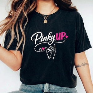 May include: Black t-shirt featuring the text "Pinky Up" in white script with pink heart accents and a hand gesture illustration. The shirt is worn with blue jeans.