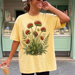 May include: A pale yellow short-sleeve t-shirt featuring a detailed botanical illustration of multiple Venus flytrap plants with red and green leaves, surrounded by green fern fronds. The shirt has a crew neckline and a loose, relaxed fit.