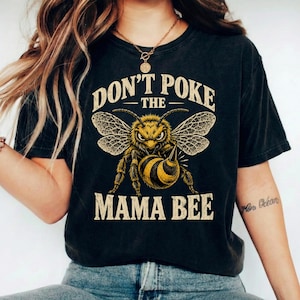 Può includere: Una t-shirt nera a maniche corte con la grafica di un'ape arrabbiata a strisce gialle e nere. Il testo sopra l'ape recita "DON'T POKE THE" e il testo sottostante "MAMA BEE" in un carattere beige dallo stile vintage e consumato.