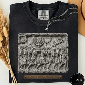 Könnte beinhalten: Ein schwarzes T-Shirt mit Rundhalsausschnitt und einem Grafikdruck des Titusbogens-Reliefs, das Figuren mit einer Menora zeigt. Das Shirt hat ein Comfort Colors-Etikett am Hals und eine silberne Stern-Halskette auf dem Stoff.