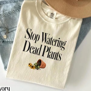 Puede incluir: Camiseta de color marfil con el texto negro "Stop Watering Dead Plants". La camiseta presenta un gráfico de una maceta de terracota volcada con girasoles muertos. Un sombrero beige y unos vaqueros azul claro están en el fondo.