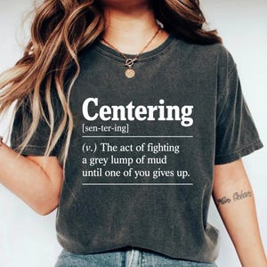 Può includere: T-shirt grigio scuro con la parola "Centering" in bianco e una definizione. La definizione recita: "(v.) L'atto di combattere un grumo grigio di fango finché uno di voi non si arrende."