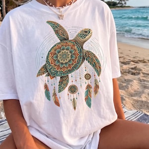 May include: A white short-sleeve t-shirt featuring a large graphic of a sea turtle with a mandala-patterned shell. The turtle is surrounded by circular line art and hangs several dangling dreamcatcher-style ornaments with feathers and beads.