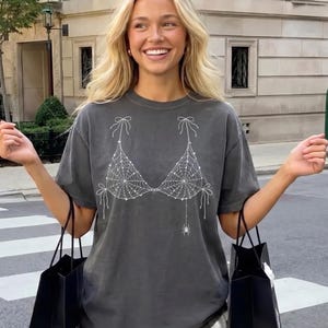 May include: A charcoal grey short-sleeve t-shirt featuring a white graphic print of a bikini top made of spiderwebs with bow details at the straps and a small spider hanging from the right side. The shirt has a crew neckline and a relaxed fit.