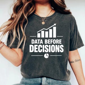 May include: A charcoal grey short-sleeve t-shirt featuring a white graphic of a bar chart with an upward trend arrow, the text "DATA BEFORE DECISIONS" in bold capital letters, and a small pie chart icon centred below the text.