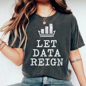 Może przedstawiać: Ciemnoszary t-shirt z krótkim rękawem z białą grafiką wykresu słupkowego w kształcie korony nad napisem LET DATA REIGN wykonanym przetartą, postarzaną czcionką. Koszulka posiada okrągły dekolt oraz wykończenie w stylu vintage.