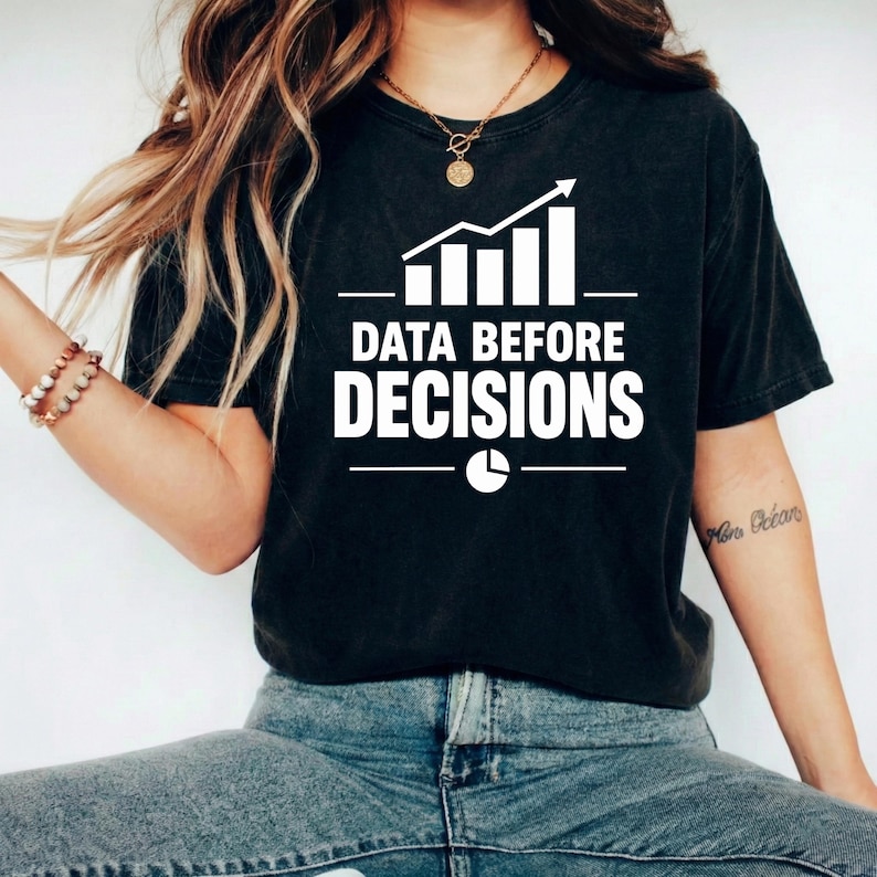 Op de afbeelding: Een zwart t-shirt met korte mouwen voorzien van een witte afbeelding van een staafdiagram met een stijgende trendpijl, de tekst "DATA BEFORE DECISIONS" in dikgedrukte hoofdletters en een klein cirkeldiagram-icoon onder de tekst op de borst.