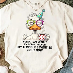 May include: White t-shirt featuring a grumpy cartoon duck wearing glasses and a party hat, holding a coffee cup. The text reads, "Sorry, I am cranky I'm going through my terrible seventies right now."