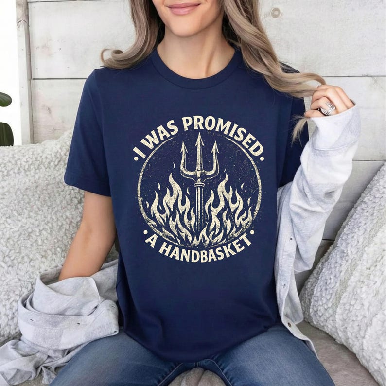 May include: Navy blue t-shirt with a distressed graphic. The graphic features a trident surrounded by flames within a circle, with the text "I WAS PROMISED. A HANDBASKET."