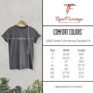 Op de afbeelding: Een donkergrijs t-shirt met korte mouwen aan een houten kledinghanger. De afbeelding bevat een maattabel met de merknaam TigroFlamingo en Comfort Colors, met afmetingen in inches voor de maten S tot 4XL, inclusief breedte en lengte.