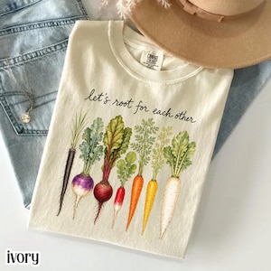 Könnte beinhalten: Ein cremefarbenes T-Shirt mit einer Grafik verschiedener bunter Wurzelgemüse, darunter Karotten, Radieschen und Rüben. Der Text "let's root for each other" ist in schwarzer Schreibschrift über der Gemüseillustration aufgedruckt.