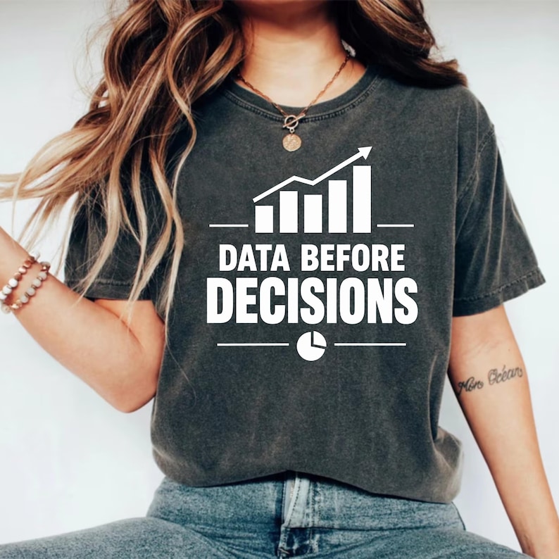 Op de afbeelding: Een antracietgrijs t-shirt met korte mouwen voorzien van een witte afbeelding van een staafdiagram met een stijgende trendpijl, de tekst "DATA BEFORE DECISIONS" in vette hoofdletters en een klein cirkeldiagram-icoon onder de tekst.