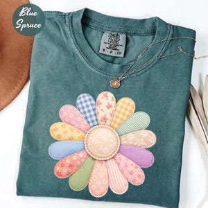 以下が含まれることがあります： 大きなカラフルなデイジーのアップリケが付いた、ティール色のTシャツ。 デイジーの花びらは、ギンガム、花柄、ストライプのデザインなど、さまざまな柄の生地で作られています。 小さなペンダントが付いたシルバーのネックレスがシャツの上にあります。 左上には「Blue Spruce」の文字があります。