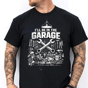 May include: Black t-shirt with a graphic design featuring the text "I'LL BE IN THE GARAGE" above crossed wrenches and various mechanic tools. The design includes a car, tools, and a coffee mug.