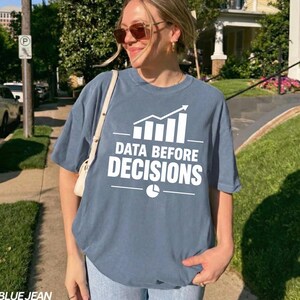 Op de afbeelding: Een leiblauw t-shirt met korte mouwen en een witte afbeelding van een staafdiagram met een pijl omhoog en de tekst "DATA BEFORE DECISIONS" boven een cirkeldiagram-icoon. Het shirt heeft een ronde hals en een relaxte pasvorm van katoen.