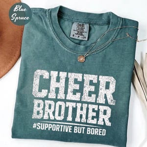 Op de afbeelding: Een teal Comfort Colors t-shirt met de woorden "CHEER BROTHER" in verweerde witte letters. Onder de tekst staat de zin "#SUPPORTIVE BUT BORED". Een gouden ketting met een ronde hanger ligt op het shirt.