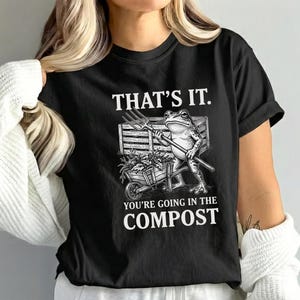 Op de afbeelding: Zwart t-shirt met een grappige afbeelding van een kikker met een hooivork, staand bij een kruiwagen vol planten. De tekst op het shirt luidt: "THAT'S IT. YOU'RE GOING IN THE COMPOST."