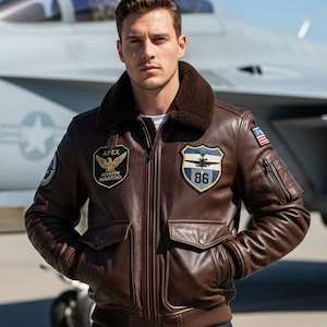 Puede incluir: Chaqueta de aviador de cuero marrón con cuello de borrego, múltiples bolsillos y parches bordados. Cuenta con cierre de cremallera y un parche de la bandera estadounidense en la manga. La chaqueta tiene el texto "APEX Aviator Squadron".