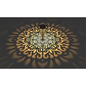 May include: A solar-powered garden stake with an ornate, gold-colored metal design. The stake casts a decorative, mandala-like pattern of light and shadow on the surface below, creating a warm, inviting ambiance.